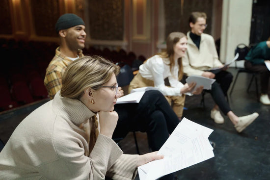 A young blonde woman in glasses and a tan turtleneck listens carefully to her teacher. She holds papers for her acting class in her other hand. In the background, several other acting students sit beside her, laughing. They are on a stage in a theater.